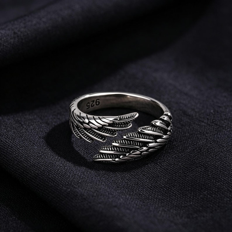 May include: A silver ring with a wing design. The ring has detailed feather patterns and the number "925" is visible. The ring is set against a dark background, highlighting its metallic sheen.