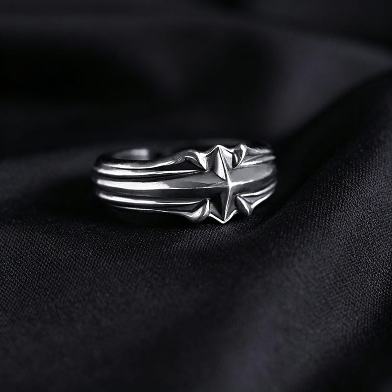 Peut inclure: Une bague en m&eacute;tal argent&eacute; pr&eacute;sentant un motif d'&eacute;toile &agrave; quatre branches pro&eacute;minent au centre. L'anneau est caract&eacute;ris&eacute; par des cr&ecirc;tes parall&egrave;les en relief qui s'&eacute;tendent horizontalement depuis le motif central, cr&eacute;ant un aspect structur&eacute;.