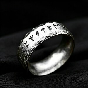 Peut inclure: Une bague en m&eacute;tal argent&eacute; pr&eacute;sentant une surface martel&eacute;e et textur&eacute;e avec des caract&egrave;res runiques grav&eacute;s sur la bande ext&eacute;rieure. La bague poss&egrave;de une finition oxyd&eacute;e sombre dans les rainures des runes et sur la texture ext&eacute;rieure rustique.