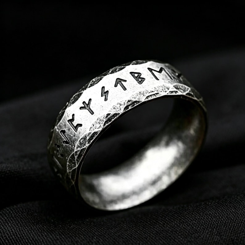 Peut inclure: Une bague en m&eacute;tal argent&eacute; pr&eacute;sentant une surface martel&eacute;e et textur&eacute;e avec des caract&egrave;res runiques grav&eacute;s sur la bande ext&eacute;rieure. La bague poss&egrave;de une finition oxyd&eacute;e sombre dans les rainures des runes et sur la texture ext&eacute;rieure rustique.