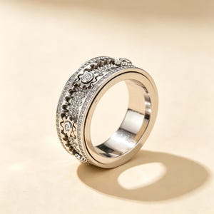 May include: A silver-toned ring featuring a wide band with a mechanical gear design. The surface is encrusted with numerous small, clear stones, and each gear element is accented with a single, larger, centered clear stone for added detail.