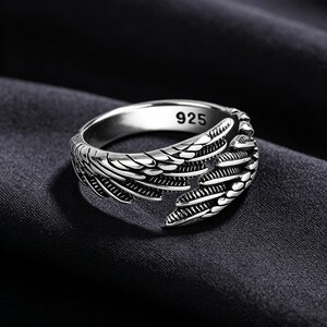 May include: A silver ring with a wing design. The ring is made of a shiny metal and features intricate feather detailing. The inside of the ring is stamped with "925". The ring is set against a dark background.