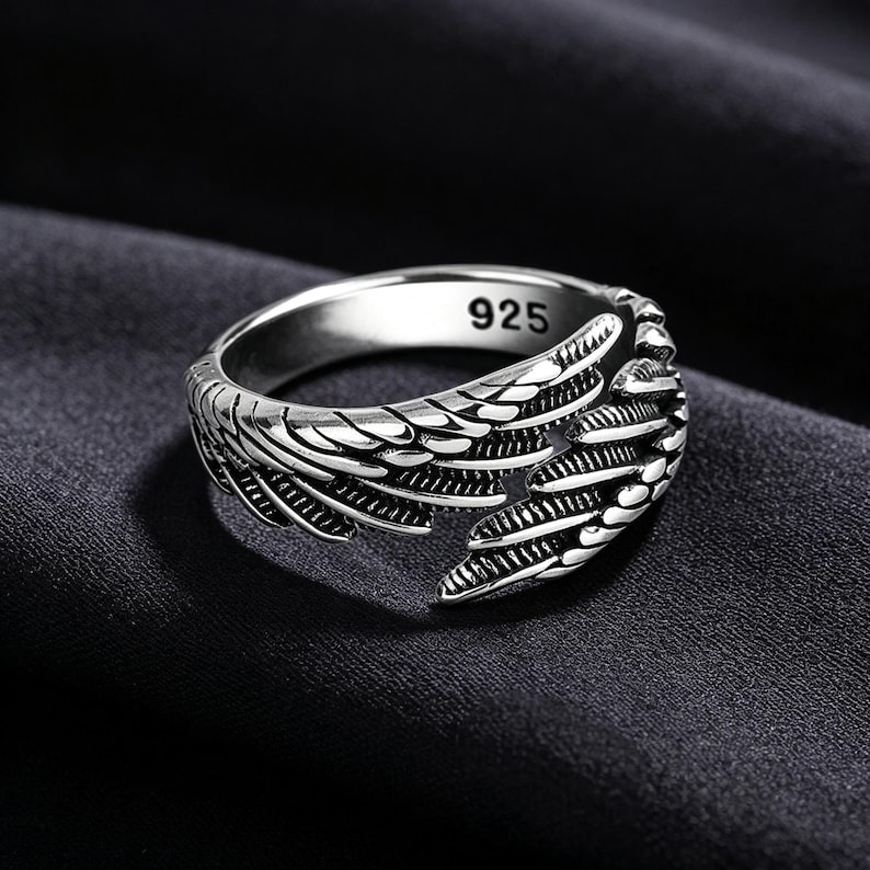 May include: A silver ring with a wing design. The ring is made of a shiny metal and features intricate feather detailing. The inside of the ring is stamped with "925". The ring is set against a dark background.