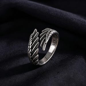 May include: A silver ring with an open wing design. The ring has a textured surface and the number "925" is visible on the band. The ring is set against a dark background. The ring is made of silver.