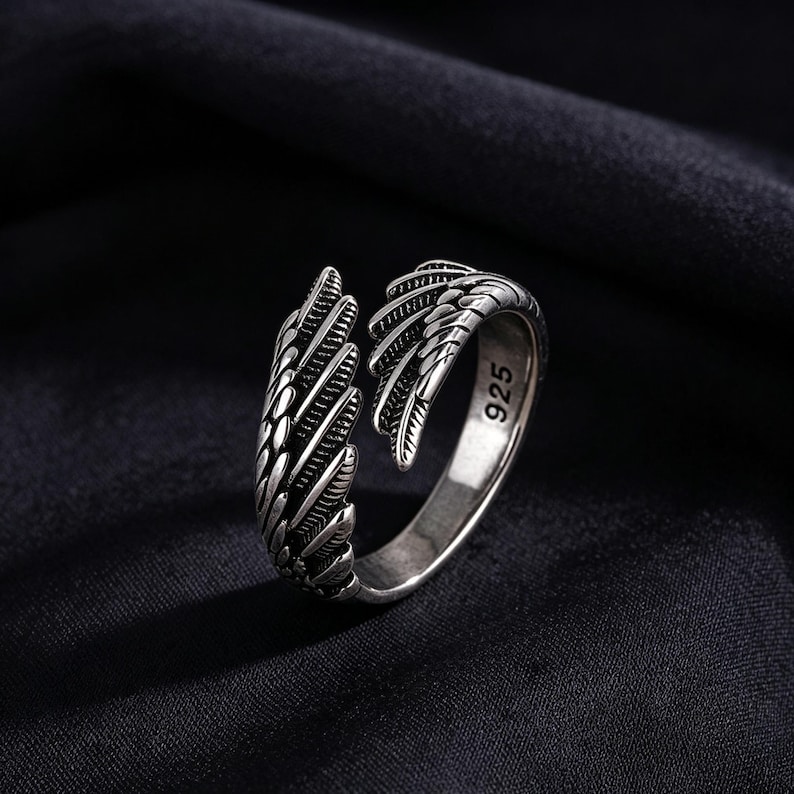 May include: A silver ring with an open wing design. The ring has a textured surface and the number "925" is visible on the band. The ring is set against a dark background. The ring is made of silver.