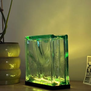 Può includere: Una lampada a blocchi di vetro verde con base nera. La lampada è illuminata da una luce soffusa e calda, creando un'atmosfera accogliente. Il blocco di vetro ha una superficie strutturata ed è posto su una superficie di legno.