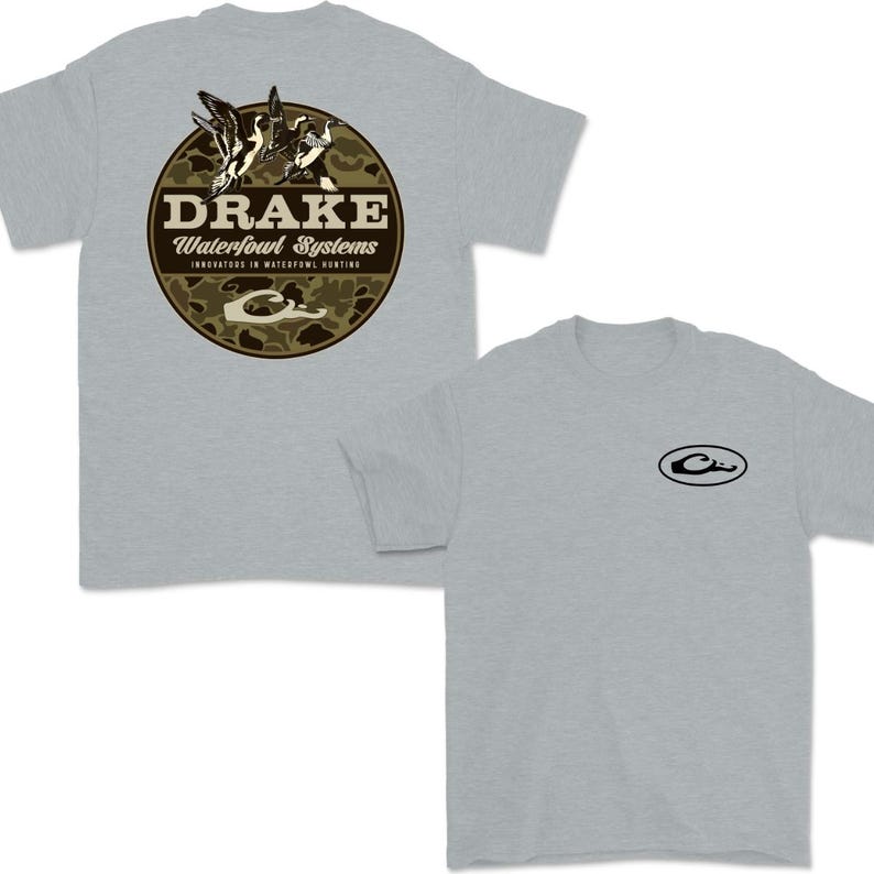 May include: Light gray t-shirt with a circular graphic on the back featuring the text "DRAKE Waterfowl Systems" and a camouflage pattern. The front of the shirt has a small oval logo. The shirt is short-sleeved.