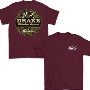 May include: Two maroon t-shirts. One features a circular design with the text "DRAKE Waterfowl Systems" and a duck illustration on a camouflage background. The other shirt has a white logo on the chest.