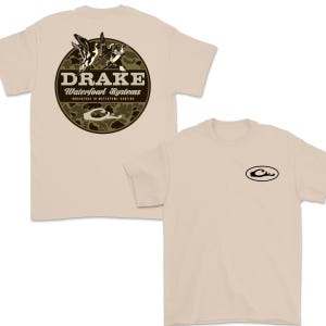 May include: Tan t-shirt featuring a circular graphic on the back with the word "DRAKE" above "Waterfowl Systems" and a camouflage pattern. The front of the shirt has a small black oval logo. The shirt is short-sleeved.