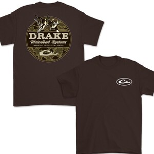 May include: Two brown t-shirts. One features a circular design with the text "DRAKE Waterfowl Systems" and a camouflage background with ducks. The other shirt has a white logo on the chest. Both shirts are short-sleeved.
