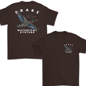May include: Two brown t-shirts with a graphic of a duck in flight. The word "DRAKE" is above the duck, and "WATERFOWL SYSTEMS" is below it. The second shirt has a smaller version of the graphic on the chest.