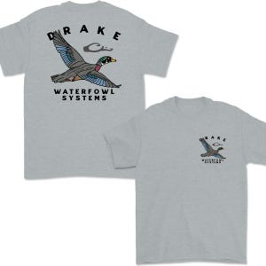 May include: Two light gray t-shirts with a duck graphic. The graphic includes a flying duck with blue, green, and red accents, the word "DRAKE" above the duck, and "WATERFOWL SYSTEMS" below. One shirt has a large graphic, the other a smaller one.