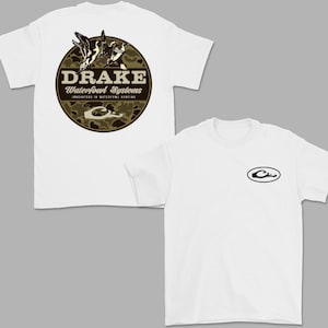 May include: Two white t-shirts. One shirt features a circular design on the back with the text "DRAKE Waterfowl Systems" and a camouflage pattern. The other shirt has a small black logo on the front.