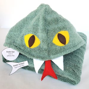 Peut inclure: Une serviette à capuche vert sauge représentant un serpent avec des yeux en feutrine jaune et marron, des crocs en feutrine blanche et une langue en feutrine rouge. Deux étiquettes en papier indiquent "Snake Eye Cut 2 yellow felt" et "Snake Tongue Cut 2 red felt."
