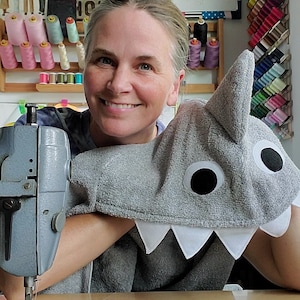 May include: A gray hooded towel featuring a shark design with a dorsal fin, large white and black circular eyes, and white triangular teeth along the bottom edge. The towel is made of a textured fabric and is positioned next to a gray sewing machine.