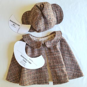 May include: A brown tweed detective hat and cape set. The hat features ear flaps and a brown ribbon. The cape has a rounded collar. Paper patterns for the hat bill and cape collar are included. The set is made of a brown and beige checked fabric.