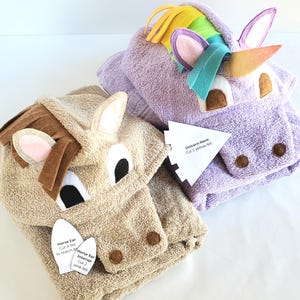 May include: A tan horse hooded towel and a purple unicorn hooded towel with a rainbow mane and horn. Both towels feature felt animal faces and ears. White paper labels are attached, with text reading Horse Ear, Horse Ear Interior, and Unicorn Horn.
