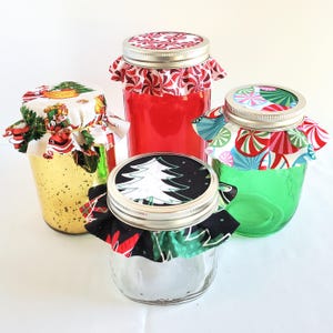 May include: Four glass jars with metal lids, each topped with decorative fabric covers. One jar is gold, one is red, one is green, and one is clear. The fabric patterns feature Christmas trees, peppermint swirls, and Santa Claus motifs.