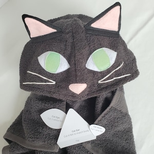 May include: A dark grey hooded towel featuring a cat face design with light green eyes, a pink nose, and white whiskers. The hood includes two pointed ears with pink centres. Attached paper labels read Cat Eye, Cat Ear, and Cat Nose.