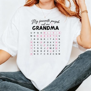 May include: A white t-shirt featuring the black text My favorite people call me GRANDMA with two small pink hearts. Below the text is a word search puzzle grid with the names Olivia, Roberta, Amber, David, and Alex highlighted in pink boxes.