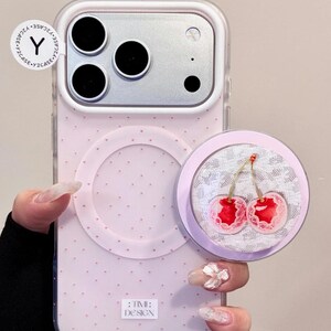 May include: Pink phone case with a cherry design pop socket. The case has a dotted pattern and a clear camera lens protector. The pop socket features two red cherries with a lace background. The text ":TIME: DESIGN" is visible.