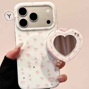 Könnte beinhalten: Weißes Handycase mit rosa Blumenmuster und herzförmigem Spiegelaufsatz. Das Case hat einen Kameraausschnitt und ein kleines rosa Herz mit dem Wort "Flur". Der Spiegel ist transparent mit einem rosa Rand.