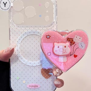 Puede incluir: Funda de teléfono transparente con un pop-socket rosa en forma de corazón con una ilustración de cerdo. La funda tiene un patrón de lunares blancos y pequeños acentos de estrellas y corazones. El pop-socket tiene una ilustración de cerdo con globos.