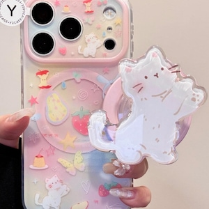 May include: Clear phone case with a pastel rainbow gradient, adorned with cartoon cat, fruit, and star charms. A white cat-shaped phone grip is attached. The case has a pink border and a clear back.