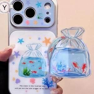 May include: A phone case with a fishbowl design. The case features a clear plastic overlay with a fishbowl graphic containing red fish, seaweed, and bubbles. The case also has a matching fishbowl charm. The text on the case reads: "The ocean is the inverted sky, and you are the sole trigger of my tears."