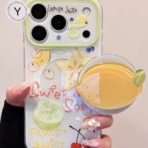 May include: Clear phone case with a lemon theme. The case features a yellow lemon slice pop socket, and illustrations of lemons, a star fruit, and the words "Lemon Juice", "Sweet & Sour", and "Fruit Addict".