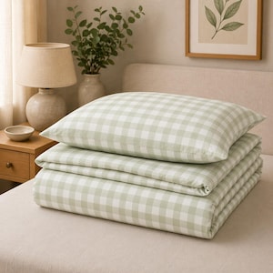 May include: A folded set of bedding with a pillow and a comforter. The set features a light green and white gingham pattern. The bedding is neatly stacked on a bed, creating a cozy and inviting aesthetic.