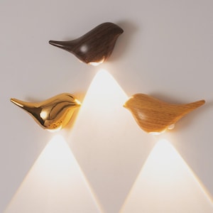 May include: Three decorative bird-shaped wall lights. One is dark brown, one is gold, and one is light brown. Each bird has a small light at the bottom, casting a warm glow. The lights are mounted on a white wall.
