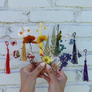 May include: A collection of clear, heart-shaped bookmarks featuring pressed dried flowers and ferns in various colors, including red, yellow, green, and blue. Each bookmark is finished with a matching colored silk tassel attached to the top loop.