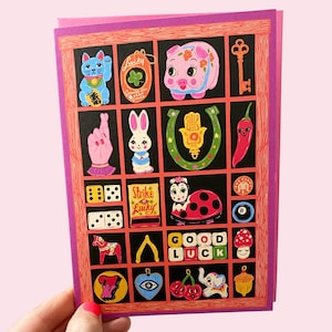 May include: A greeting card with a pink border and background, featuring a grid of lucky charms. The charms include a waving cat, four-leaf clover, pig, key, rabbit, horseshoe, chili pepper, dice, ladybug, and mushroom.