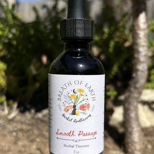 May include: A black glass bottle with a dropper, labeled "Breath of Earth Herbal Apothecary." The label features a floral illustration and the words "Smooth Passage Herbal Tincture." The bottle contains 2 oz of liquid and sits on a rock.