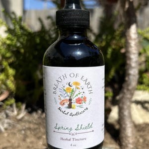 Puede incluir: Una botella de vidrio oscuro con un cuentagotas negro. La etiqueta dice "Breath of Earth Herbal Apothecary Spring Shield Herbal Tincture 118 ml." La etiqueta presenta una ilustración floral con una luna creciente y estrellas.