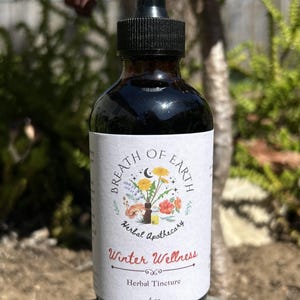 Puede incluir: Una botella de vidrio ámbar oscuro con un cuentagotas negro, etiquetada como "Breath of Earth Herbal Apothecary". La etiqueta presenta ilustraciones florales y el texto "Winter Wellness Herbal Tincture". La botella contiene 118 ml de líquido.