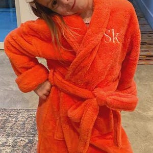 May include: A vibrant orange terrycloth robe with a matching belt and the initials "SK" embroidered on the chest. The robe is worn with white slippers. The image is taken indoors.