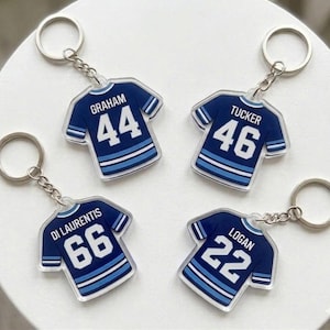 May include: Four acrylic keychains shaped like sports jerseys in dark blue with light blue stripes. Each features a white name and number: GRAHAM 44, TUCKER 46, DI LAURENTIS 66, and LOGAN 22. Each jersey is attached to a silver metal split ring by a chain.