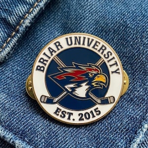 Op de afbeelding: Een ronde pin met een gouden rand met de tekst "BRIAR UNIVERSITY" in wit. Het midden heeft een blauwe achtergrond met een gestileerde adelaar en gekruiste hockeysticks. De tekst "EST. 2015" staat onderaan.