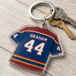 May include: A clear acrylic keychain in the shape of a football jersey. The jersey is navy blue with the name "GRAHAM" and the number "44" in white. The jersey has red and gold accents. A silver keyring holds the keychain and two keys.