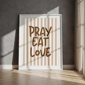 May include: A framed print with the words "PRAY EAT LOVE" in brown lettering. The background features vertical stripes in beige and off-white. The frame is white, and the print is displayed against a gray wall.