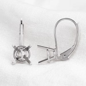 May include: Two silver-colored earring findings. One has a round setting with four prongs, and the other has a lever-back closure. The findings are displayed on a white, textured surface.