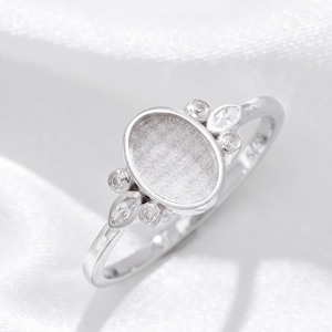 Könnte beinhalten: Ein silberfarbener Ring mit einer zentralen, ovalen Fassung, die eine texturierte, mattierte Innenseite aufweist. Die Fassung ist mit vier kleinen, klaren, marquise- und rundgeschliffenen Steinen verziert, die symmetrisch an den Seiten angeordnet sind.
