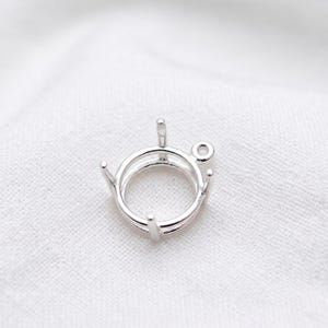 Puede incluir: Un engaste de joyería de color plateado con un diseño circular. Cuenta con un anillo para sujetar una piedra preciosa, con cuatro garras y un pequeño lazo para sujetarlo a una cadena u otro componente de joyería. El engaste está sobre un fondo blanco.