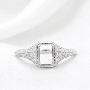 May include: A silver-toned metal ring featuring an empty rectangular setting at the center. The band is decorated with intricate filigree patterns and small, clear, round stones embedded along the sides of the shank, showcasing a detailed design.