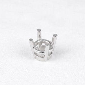May include: A silver-colored jewelry setting with four prongs extending upwards. The setting has a circular base and is designed to hold a gemstone. The metal has a polished finish, reflecting light.
