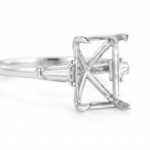Puede incluir: Un engaste de anillo de compromiso plateado con un centro rectangular y detalles laterales con corte baguette. El anillo se presenta sobre un fondo blanco, mostrando su intrincado diseño y acabado pulido.