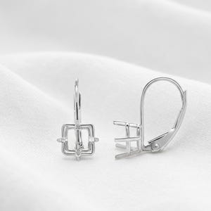 May include: A pair of silver-toned leverback earring settings featuring a square-shaped prong mounting designed to hold a gemstone. The metal components have a polished finish and a secure hinged closure mechanism for attachment to the earlobe.