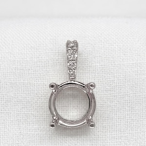 May include: A silver pendant setting with a round bezel and a bail adorned with small, sparkling stones. The pendant has four prongs and is designed to hold a round gemstone. The setting is displayed against a white background.